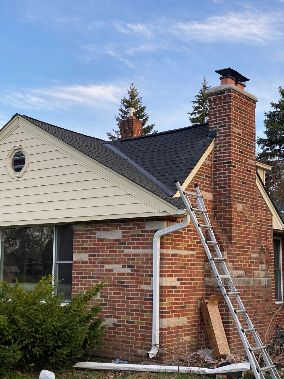 The same roof and brick chimney after restoration, with new dark shingles, a brick chimney topped with a metal cap, and the gutter and downspout in place.